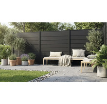 Moderner Sichtschutzzaun aus dunklen Paneelen auf einer Terrasse mit Holzbank, Steinplatten und verschiedenen Pflanzen in Töpfen.