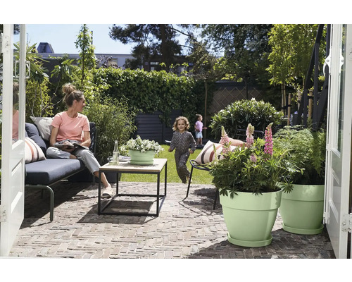Terrasse moderne avec canapé de jardin, table en bois et pots vert clair. Femme sur le canapé, enfant jouant sur la pelouse à l'arrière-plan.