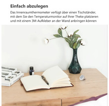 Kleines rundes Innenthermometer auf einem Holzschreibtisch neben einem Notizbuch und einer Vase mit Blumen.