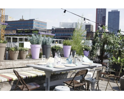 Terrasse de toit lumineuse avec table de jardin dressée, chaises pliantes, banc en bois et divers pots de fleurs devant un décor urbain.