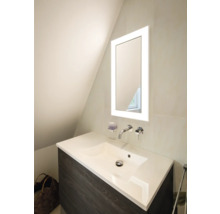 Meuble sous-vasque moderne avec lavabo blanc, mitigeur mural chromé et miroir éclairé dans une salle de bains lumineuse.