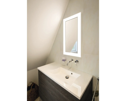 Meuble sous-vasque moderne avec lavabo blanc, mitigeur mural chromé et miroir éclairé dans une salle de bains lumineuse.