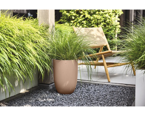 Pot de fleurs rond marron avec structure nervurée et graminées vertes sur une terrasse en gravier sombre à côté d une chaise en bois.