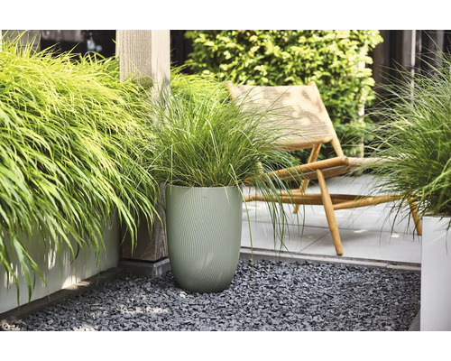 Herbe ornementale dans un grand pot de fleurs vert texturé sur une terrasse moderne avec du gravier gris et une chaise longue en bois.
