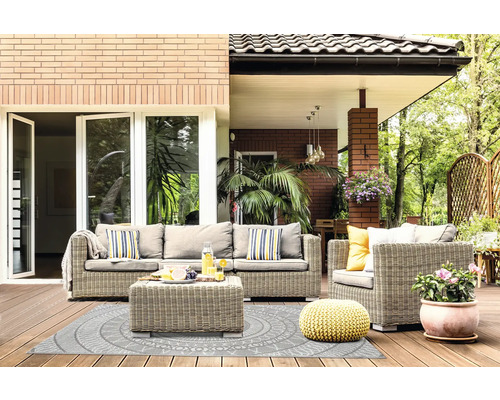 Terrasse lumineuse avec salon de jardin en rotin gris, tapis d'extérieur à motifs circulaires, pouf jaune et plante en pot sur plancher en bois.