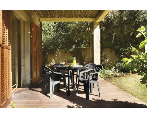 Salon de jardin avec table ronde noire et quatre fauteuils en plastique sur une terrasse en bois ensoleillée sous une pergola.