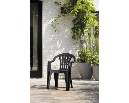 Chaise de jardin empilable en plastique noir avec accoudoirs sur une terrasse claire devant un mur en briques blanches avec des plantes.