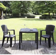 Deux chaises de jardin anthracite et une table carrée aspect rotin sur une terrasse en bois devant un jardin verdoyant avec pelouse et arbres.