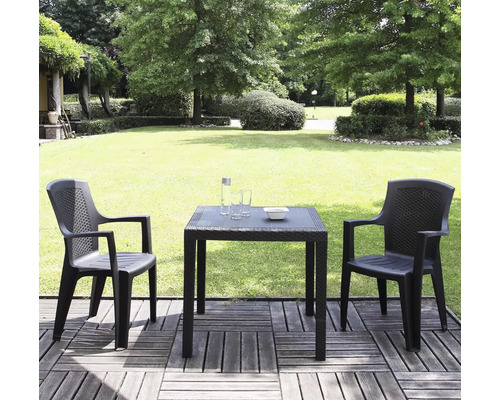 Deux chaises de jardin anthracite et une table carrée aspect rotin sur une terrasse en bois devant un jardin verdoyant avec pelouse et arbres.