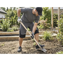 Ein Mann in grauer Arbeitskleidung und Schutzhandschuhen gräbt mit einem Spaten in einem Gartenbeet um.