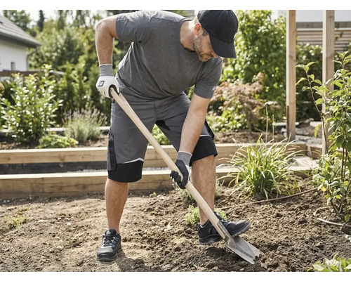 Ein Mann in grauer Arbeitskleidung und Schutzhandschuhen gräbt mit einem Spaten in einem Gartenbeet um.