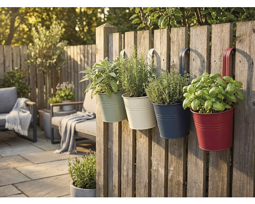 Quatre pots de fleurs suspendus colorés en métal avec des herbes sur une clôture en bois dans un jardin lumineux avec mobilier de terrasse.