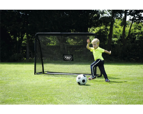 Enfant jouant au football devant un but de foot EXIT TOYS noir sur une pelouse avec des arbres en arrière-plan.