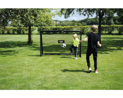 Deux enfants jouent au football avec un but de football EXIT Toys sur une pelouse dans un jardin clair et ensoleillé.