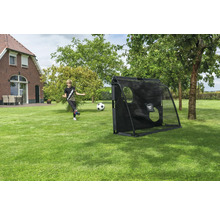 Un garçon tire un ballon vers un but de foot noir avec EXIT TOYS Logo et bâche d'entraînement dans un grand jardin avec pelouse et arbres.