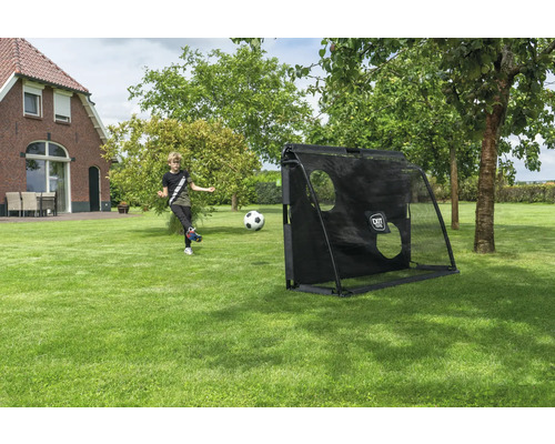 Un garçon tire un ballon vers un but de foot noir avec EXIT TOYS Logo et bâche d'entraînement dans un grand jardin avec pelouse et arbres.