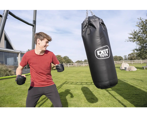 Un jeune homme avec des gants de boxe dans un jardin à côté d'un sac de frappe noir EXIT TOYS suspendu à un cadre métallique sur une pelouse.