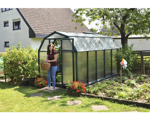 Garten-Gewächshaus mit dunkelgrünem Rahmen, Scheunenform und lichtdurchlässigen Paneelen, eine Frau mit Korb steht in der offenen Tür.
