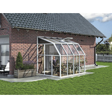 Anlehngewächshaus mit weißem Aluminiumrahmen und Glaspaneelen an einer Backsteinwand in einem Garten mit Pflanzen.