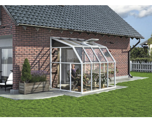 Anlehngewächshaus mit weißem Aluminiumrahmen und Glaspaneelen an einer Backsteinwand in einem Garten mit Pflanzen.