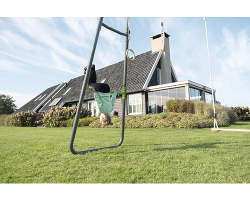 Portique de jeux en métal dans un jardin avec barre de traction, anneaux de gymnastique et balançoire, enfant la tête en bas devant une maison.