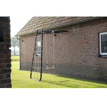 Portique de jeux mural avec structure en métal noir, balançoire en bois et filet à grimper fixé sur un mur en briques dans un jardin.
