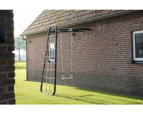Portique de jeux mural avec structure en métal noir, balançoire en bois et filet à grimper fixé sur un mur en briques dans un jardin.