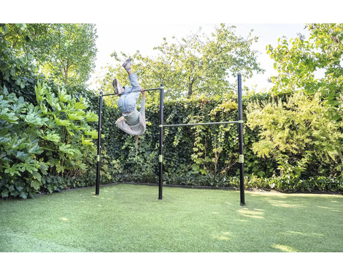 Barre de gymnastique double en métal dans le jardin, un enfant fait de la gymnastique la tête en bas, entouré de haies, atmosphère claire.