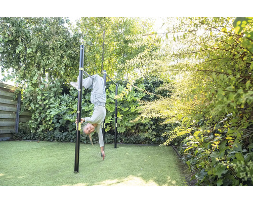 Un enfant fait de la gymnastique sur une barre fixe en métal noir dans un jardin vert et lumineux avec gazon synthétique.