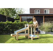 Un enfant joue avec un jeu d'eau en bois EXIT Toys dans un jardin. Le jeu comporte plusieurs rigoles et un bassin sur l'herbe. Logo EXIT Toys.