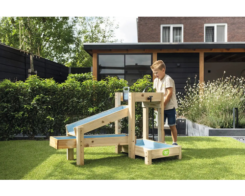 Un enfant joue avec un jeu d'eau en bois EXIT Toys dans un jardin. Le jeu comporte plusieurs rigoles et un bassin sur l'herbe. Logo EXIT Toys.