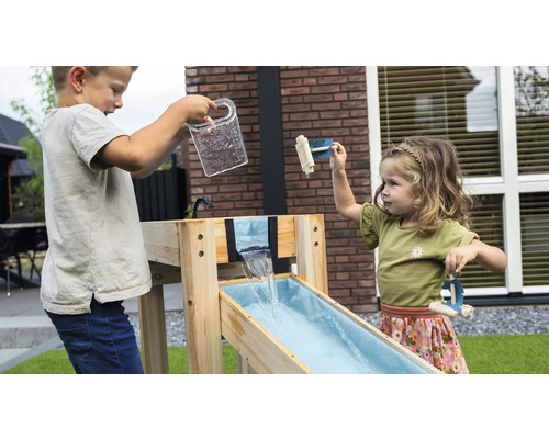 Deux enfants jouent avec un circuit d'eau en bois dans le jardin. Un garçon verse de l'eau, une fille joue avec des petits radeaux en bois.