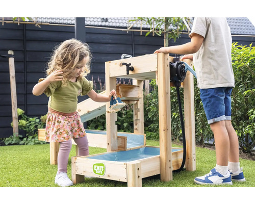 Des enfants jouent dans un jardin avec un circuit d eau EXIT Toys en bois, équipé d une pompe à eau et de plusieurs niveaux sur du gazon vert.