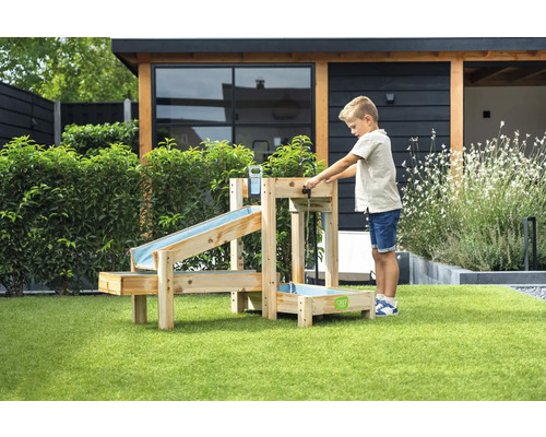 Enfant joue dans le jardin avec un circuit d'eau en bois à plusieurs niveaux et des bassins de récupération sur une pelouse devant un abri de jardin.