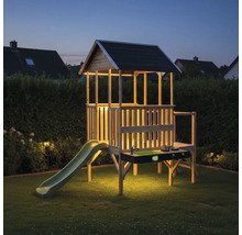 Beleuchtetes Stelzenhaus aus Holz für Kinder mit Rutsche, Dach und integrierter Spielküche im Garten bei Dämmerung.