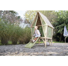 Ein Kinder-Spielhaus aus Holz mit grüner Rutsche und Leiter steht in einem sandigen Gartenbereich. Ein Mädchen sitzt oben auf der Rutsche.