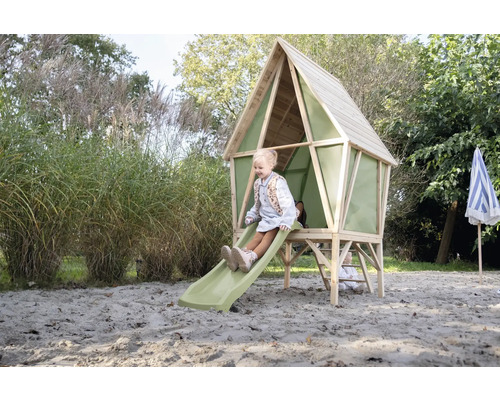 Ein Kinder-Spielhaus aus Holz mit grüner Rutsche und Leiter steht in einem sandigen Gartenbereich. Ein Mädchen sitzt oben auf der Rutsche.
