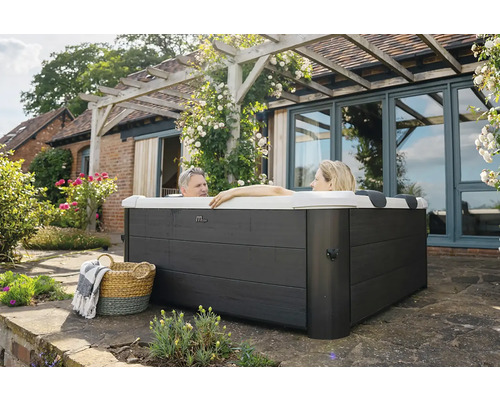 Deux personnes se détendent dans un spa extérieur carré avec habillage gris foncé sur une terrasse à côté d'une maison avec pergola.