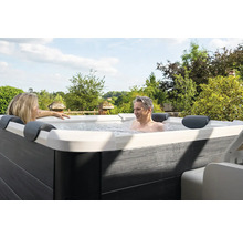 Un homme et une femme se détendent dans un jacuzzi extérieur avec un habillage gris aspect bois et une coque intérieure blanche dans un jardin.