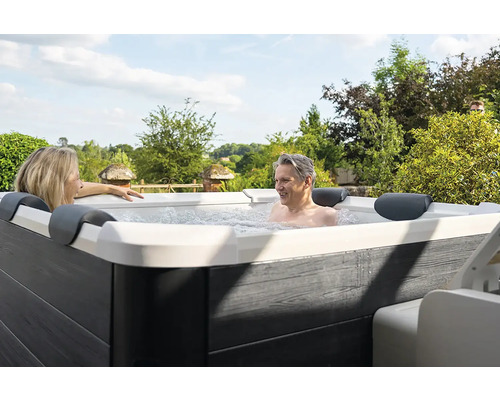Un homme et une femme se détendent dans un jacuzzi extérieur avec un habillage gris aspect bois et une coque intérieure blanche dans un jardin.