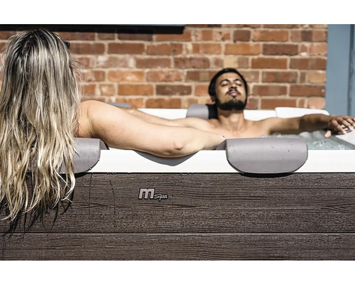 Un homme et une femme se détendent dans un jacuzzi MSpa avec un habillage aspect bois devant un mur de briques.