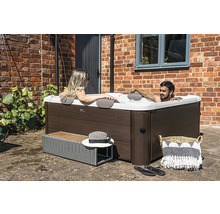 Spa carré avec habillage aspect bois, deux personnes se détendent dans l'eau sur une terrasse devant un mur en briques.