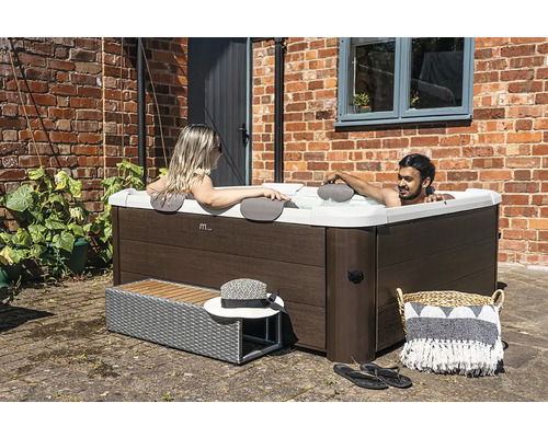 Spa carré avec habillage aspect bois, deux personnes se détendent dans l'eau sur une terrasse devant un mur en briques.