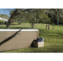 Spa MSpa avec panneaux aspect bois dans un grand jardin avec pelouse et arbres. Un panier avec des serviettes se trouve à côté.