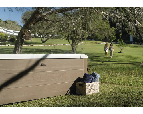 Spa MSpa avec panneaux aspect bois dans un grand jardin avec pelouse et arbres. Un panier avec des serviettes se trouve à côté.