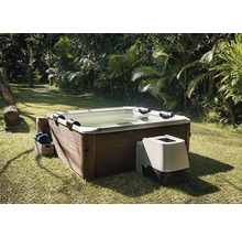 Spa carré avec habillage aspect bois et unité de pompage externe, sur une pelouse dans un jardin avec des palmiers.