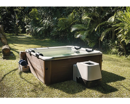 Spa carré avec habillage aspect bois et unité de pompage externe, sur une pelouse dans un jardin avec des palmiers.