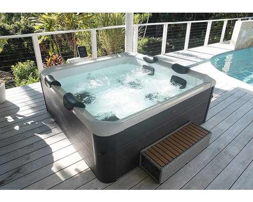 Jacuzzi extérieur avec habillage gris foncé et coque intérieure blanche, appuie-tête et eau bouillonnante sur une terrasse en bois.