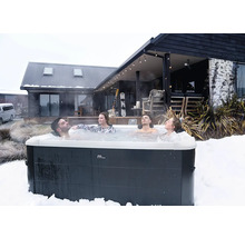 Quatre personnes se relaxent dans un spa extérieur MSpa dans un paysage enneigé devant une maison moderne en bois.