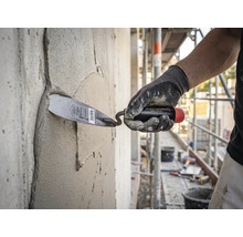 Ein Handwerker trägt mit einer Maurerkelle Mörtel auf eine Betonwand auf einer hellen und organisierten Baustelle auf.